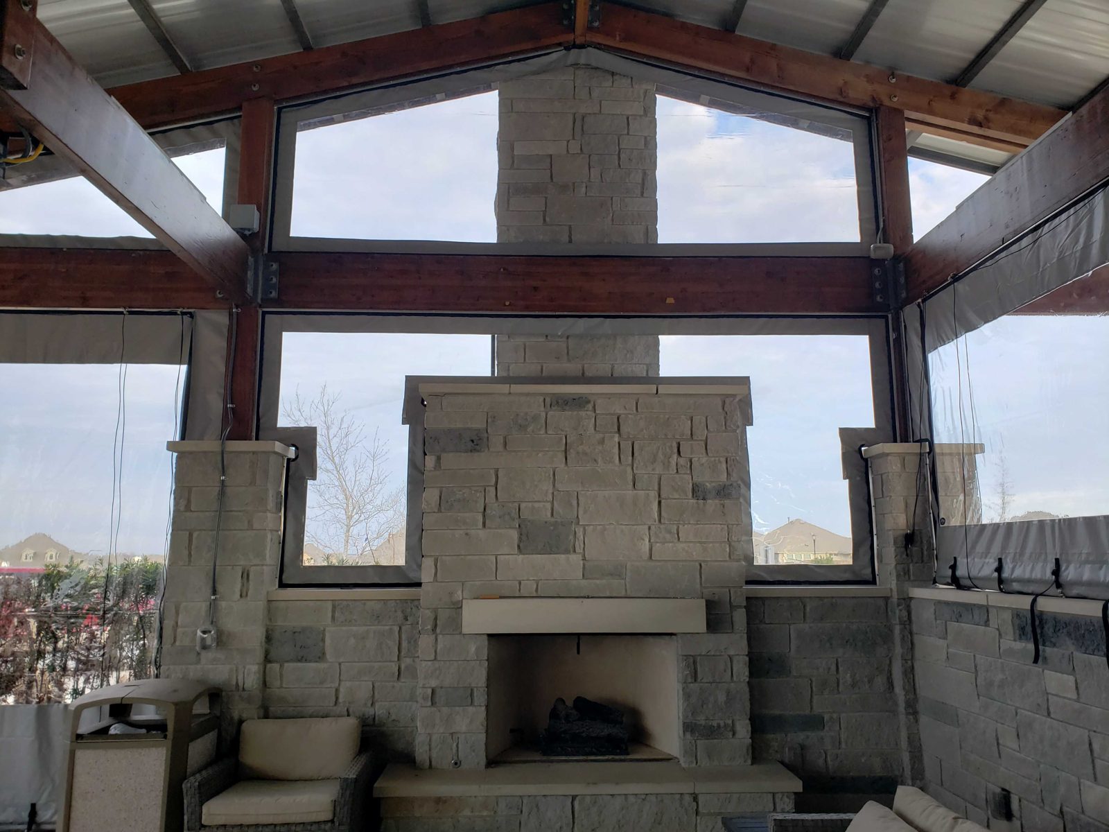 Inside the Crosswinds pavilion: clear panels filling the gable around the stone fireplace, rooftops of the neighborhood through the vinyl