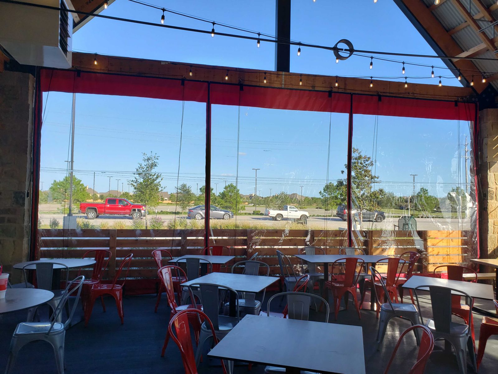 Hat Creek Fulshear from inside the patio: red-bordered clear panels down along the rail, the string lights on and the road beyond