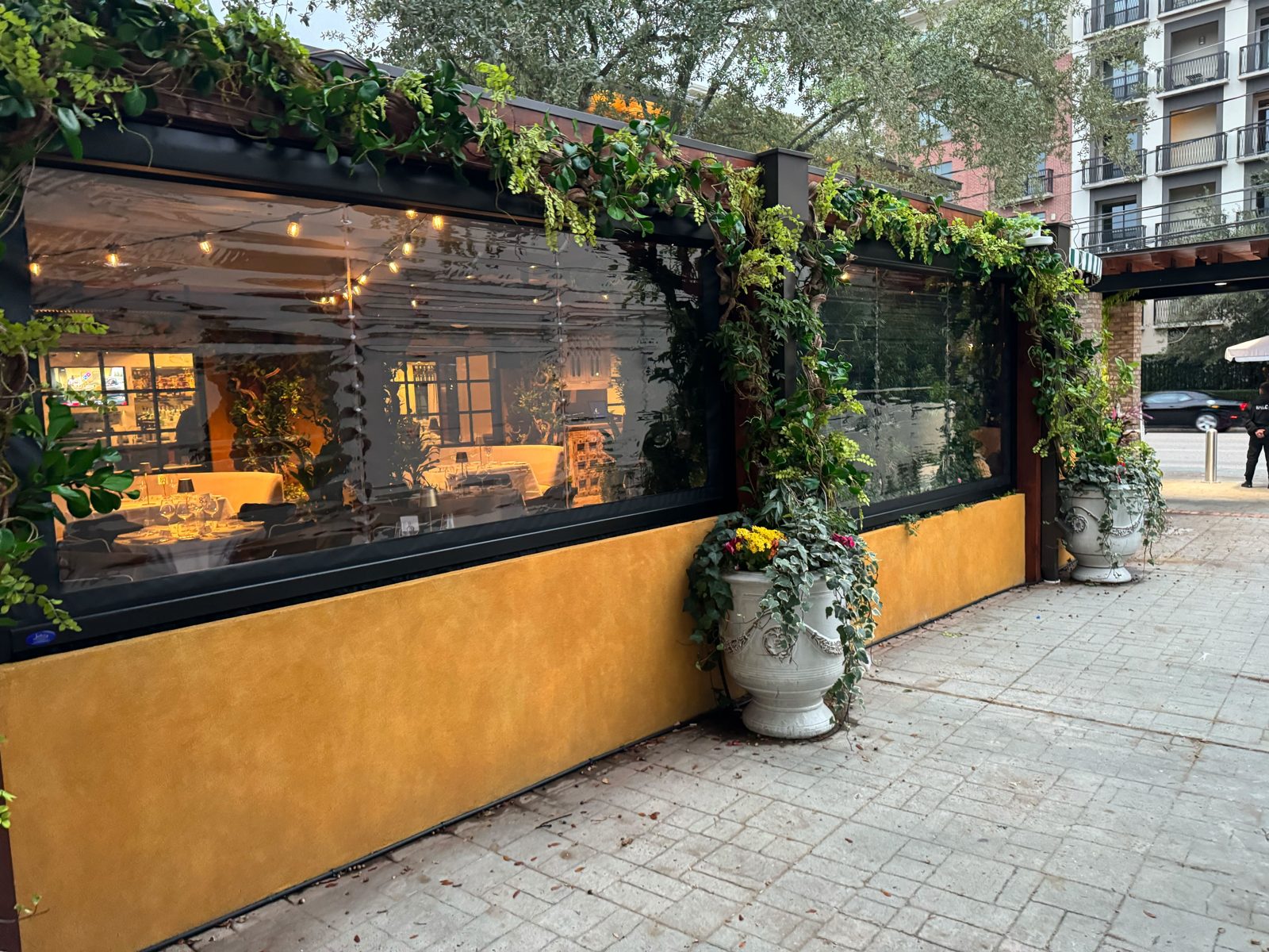 The straight run of La Griglia's patio: motorized clear screens above the yellow wall with ivy on the posts and urns at the base
