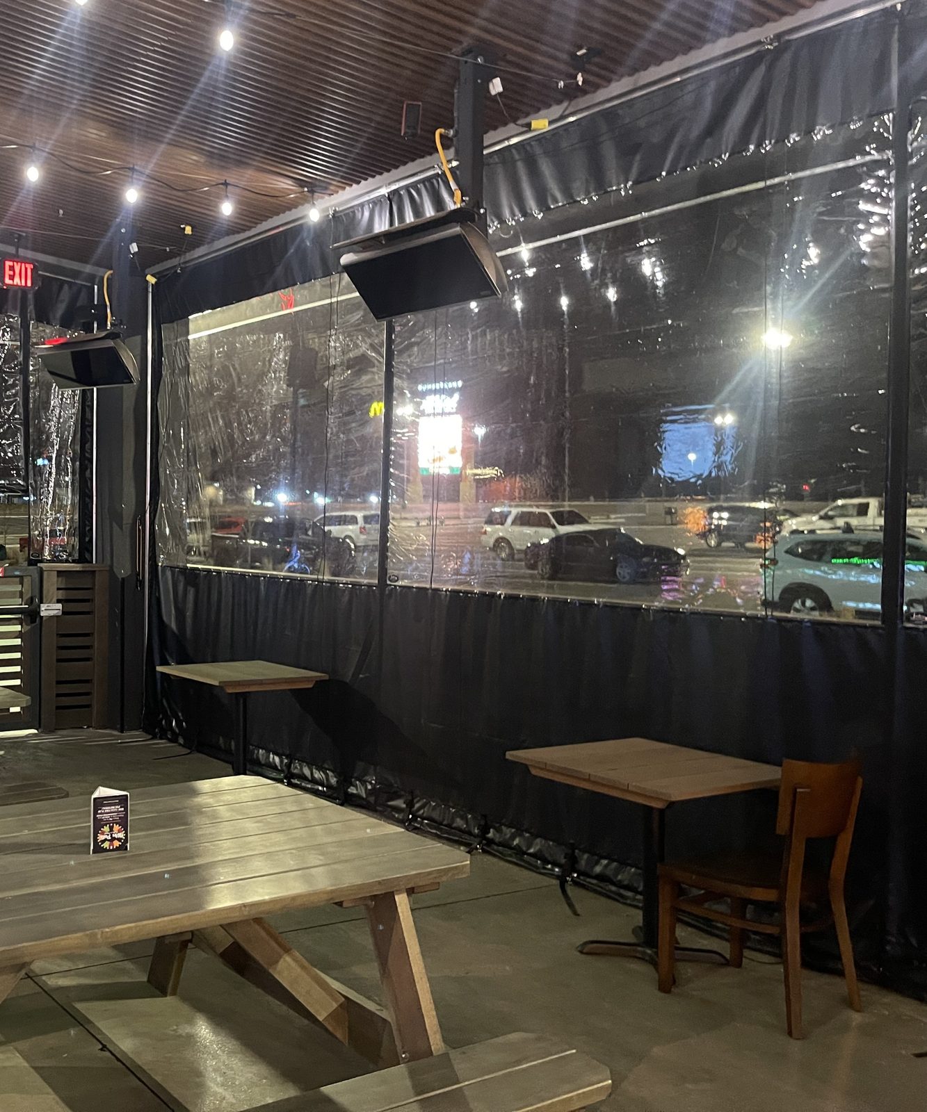 Inside The Back Yard patio at night: picnic tables, string lights and the clear panels closed against the parking lot