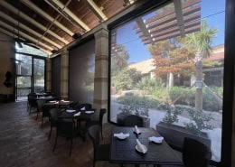 The Cheesecake Factory San Antonio – Interior View of Motorized Nano 97 Sun Shades Interior view of motorized Nano 97 black sun shades at The Cheesecake Factory in San Antonio’s La Cantera – clear view through the fabric showing hoods, side tracks, weight bar, egress door, fixed gable panel, dining tables, chairs, ceiling fans, and speakers