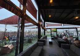Kirby Ice House Gessner Houston – Interior View of Manual Clear Vinyl Patio Enclosure Interior view of manual roll clear vinyl patio enclosure with black trim at Kirby Ice House Gessner in Houston, Texas by Southern Patio Enclosures – showing sofas, heaters, sun shades, clear curtains, fixed panels for 20 ft tall areas, and man-lift installation on this massive commercial patio