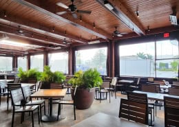 Lazy Dog Restaurant & Bar Interior view of manual roll clear vinyl patio enclosure with black trim at Lazy Dog Restaurant & Bar in Boca Raton, Florida by Southern Patio Enclosures – showing ceiling fans, heaters, plants, dining tables and chairs, and the clear panels looking out through the complex structure with beams and rock base columns