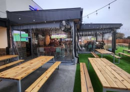Pluckers Wing Bar Austin (The Linc) – Manual Clear Vinyl Patio Enclosure (Side View) Side view of manual clear vinyl roll-down patio enclosure at Pluckers Wing Bar The Linc in Austin, Texas by Southern Patio Enclosures – showing wooden picnic tables on turf and concrete, industrial fans, string lights, and full clear panels that open the entire sports-bar patio to the outdoors