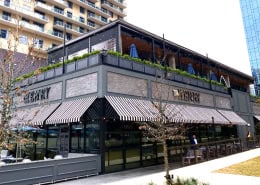 The Henry Dallas – Motorized Patio Enclosure by Southern Patio Enclosures Motorized patio enclosure on the upper-level terrace at The Henry Dallas by Southern Patio Enclosures – modern black roll-down system with striped awnings below, creating year-round outdoor dining in the heart of Dallas with high-rise views