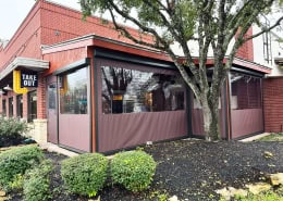 Willie’s Roadhouse Texas – Motorized Clear Vinyl Patio Enclosure Motorized clear vinyl patio enclosure with maroon solid vinyl bottom panels at Willie’s Roadhouse in Texas by Southern Patio Enclosures – clean black tracks and hardware on the red brick building with large trees and landscaping